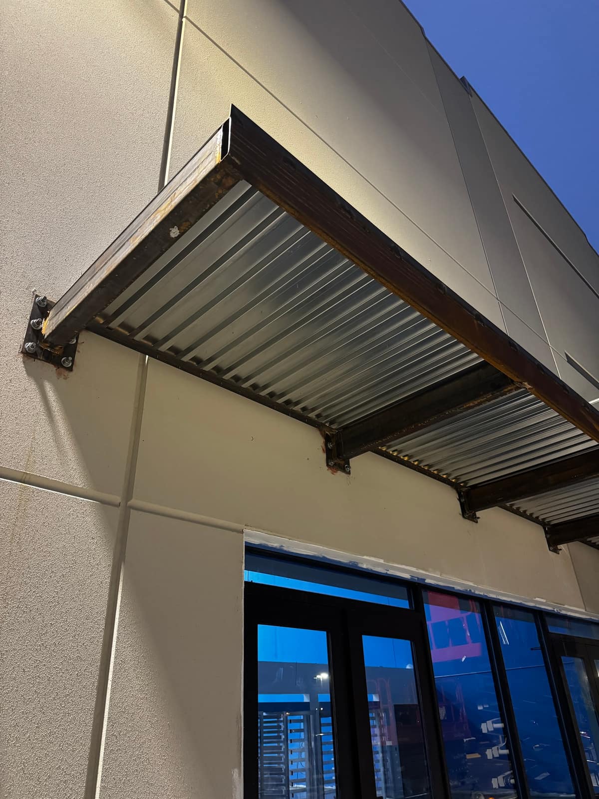 Underside of the finished steel canopy at dusk, its tube frame and ribbed metal deck ceiling cantilevered off the wall above a glass entrance